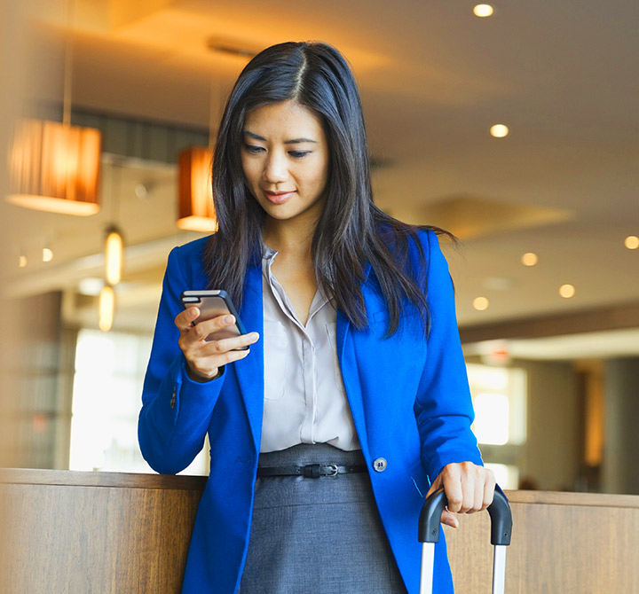Business woman holding a smartphone in one hand and a roller suitcase in another hand.